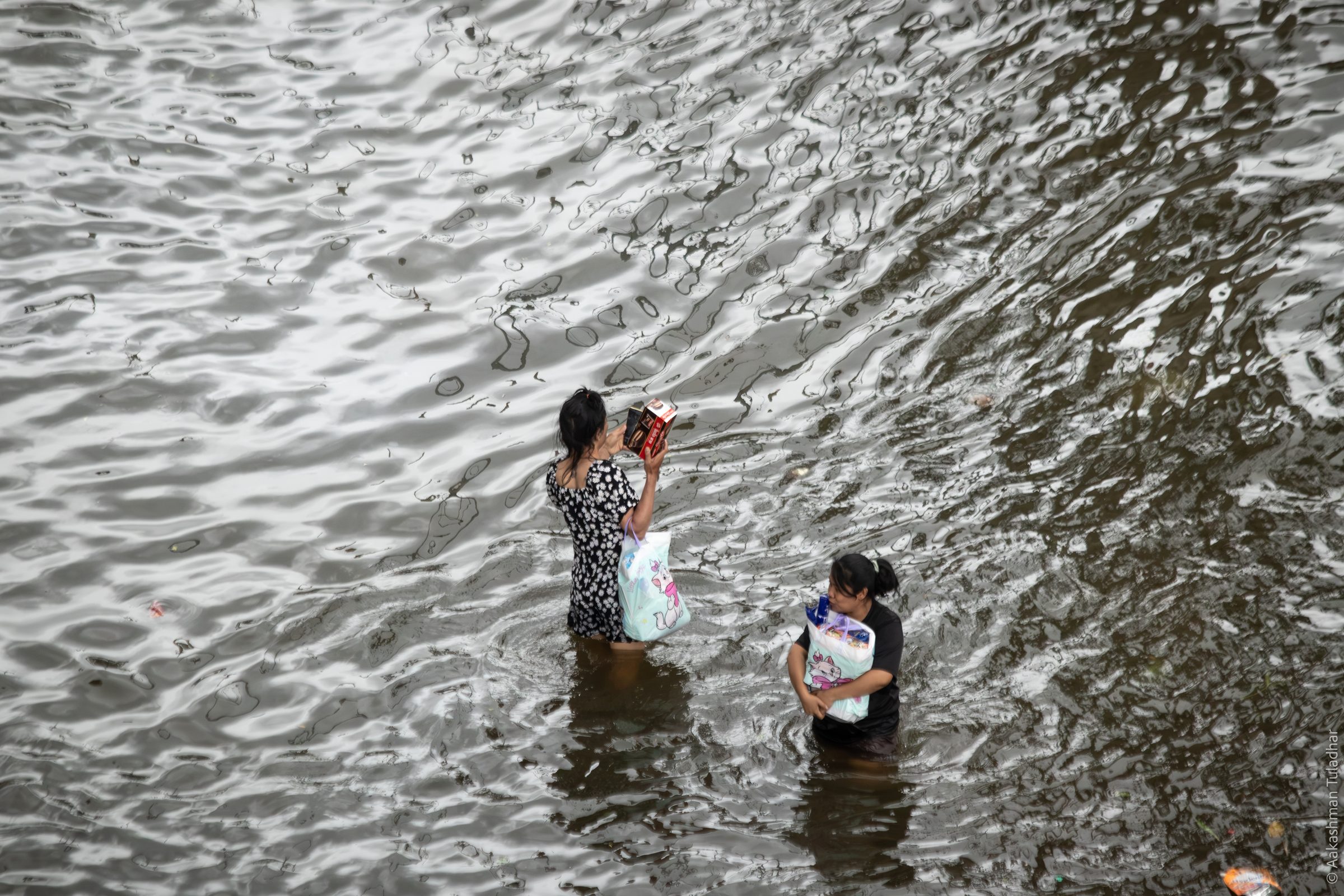 Two women wading through rippling floodwater carrying shopping bags, seen from above