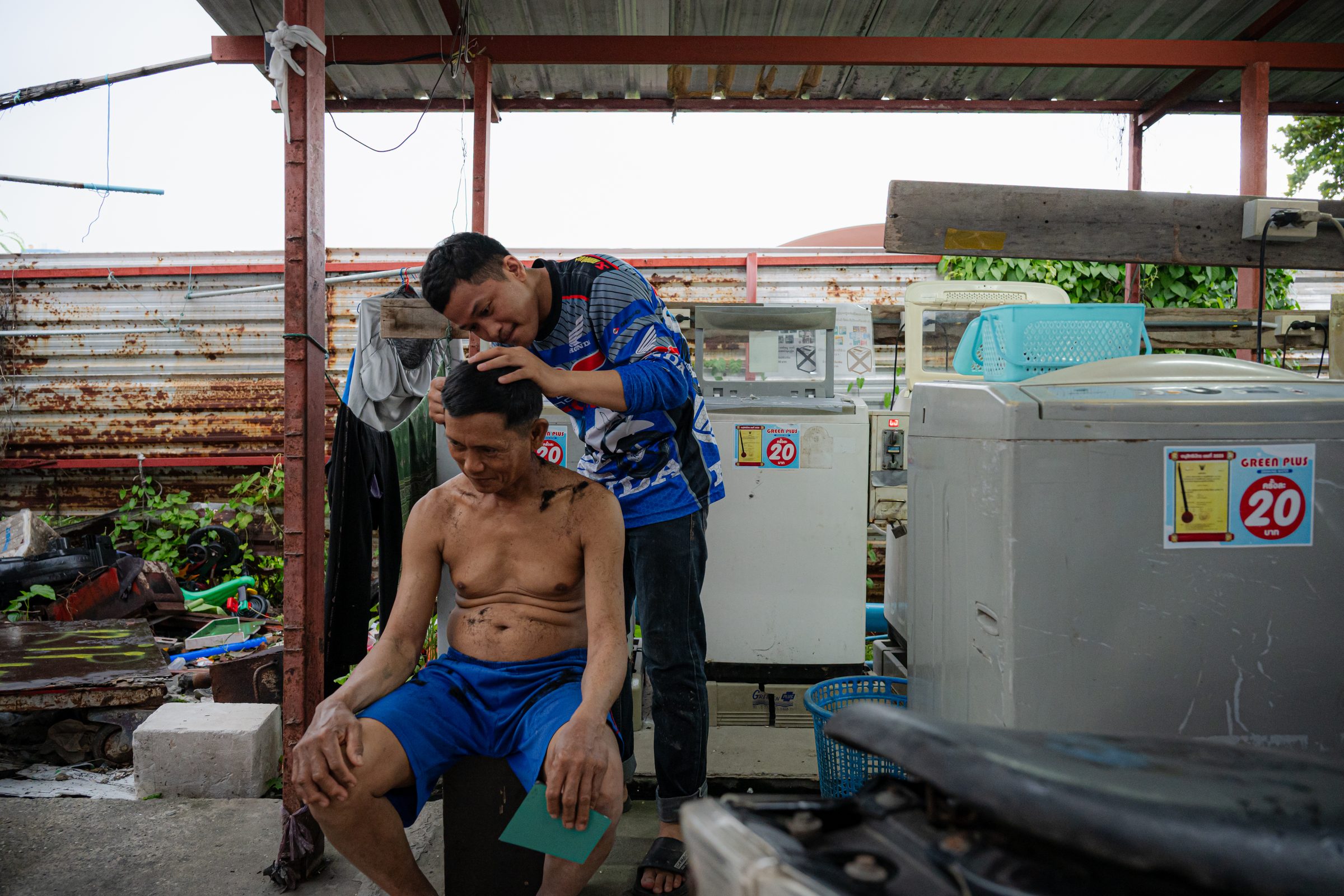 Man cutting another man's hair under a tin-roofed shelter beside washing machines