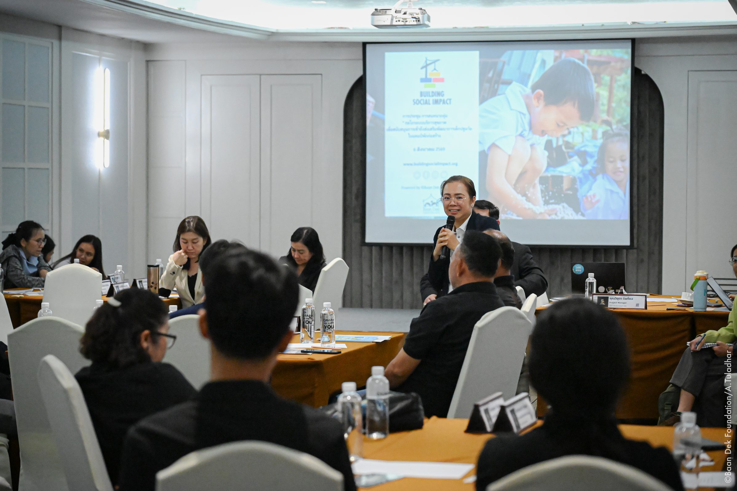 Woman standing with a microphone beside a projected Building Social Impact slide at a partnerships meeting