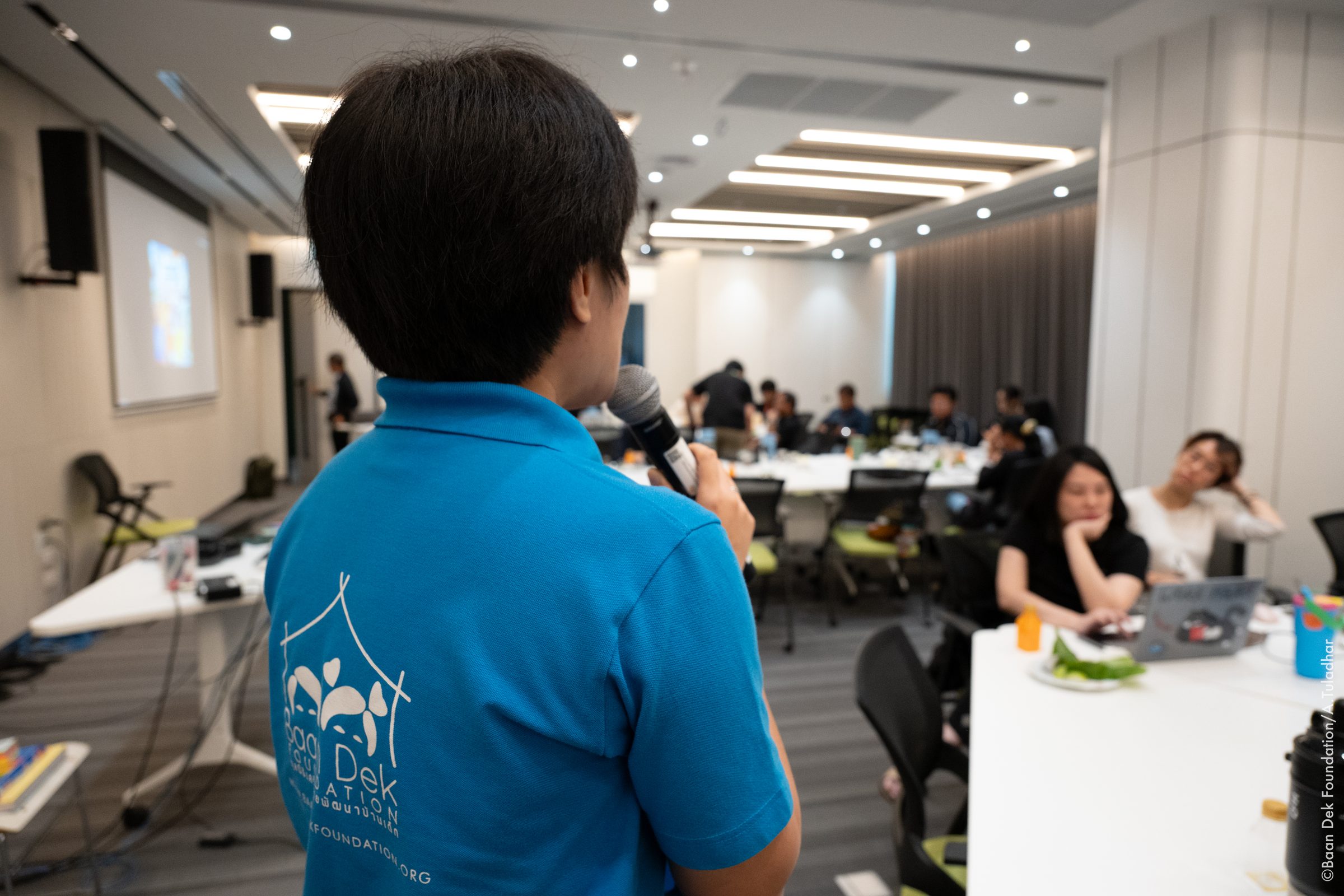 Staff member in a Baan Dek Foundation shirt speaking into a microphone at a workshop, seen from behind