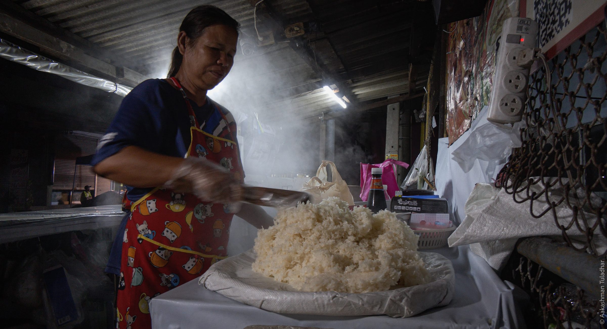 Woman handling a large mound of steaming rice at a food stall under a corrugated roof